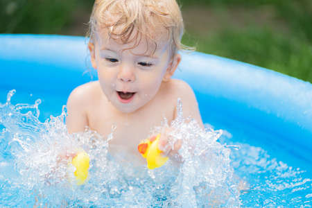 Cute little child bathing with duck in blue street pool in courtyard. Portrait of joyful toddler, baby. Kid laughs, splashes water, smiles. Concept of healthy lifestyle, family, leisure in summerの写真素材