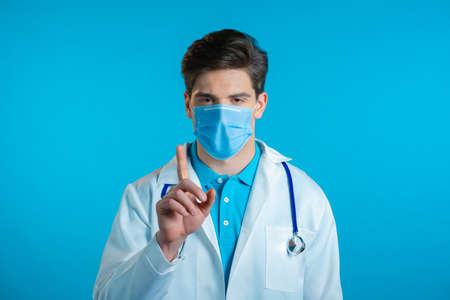 Portrait of serious doctor in professional medical white coat and mask showing rejecting gesture disapproving with no finger sign. Denying, Disagree. Doc man isolated on blue background.の写真素材
