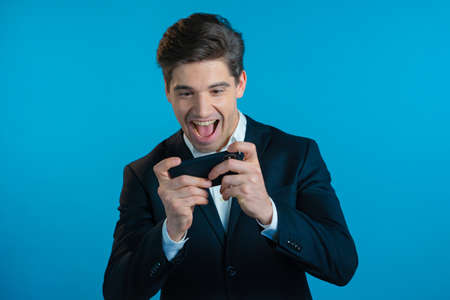 Handsome business man playing game on smartphone on blue studio wall. Using modern technology -...