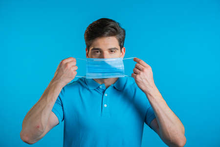 Young man puts on face medical mask during coronavirus pandemic. Portrait on blue background. Protection with respirator against COVID-19 outbreakの写真素材