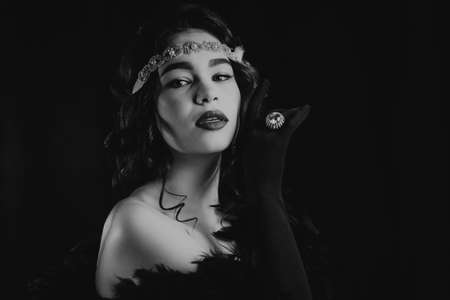 Black and white portrait of lady dressed in roaring twenties era with feathers boa on dark background. Retro, party, fashion concept. Sepia film effect.の写真素材