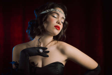 Portrait of old-fashioned woman dressed in Art deco era flirting and posing on burgundy velours background. Vintage, flapper party, retro fashion conceptの写真素材