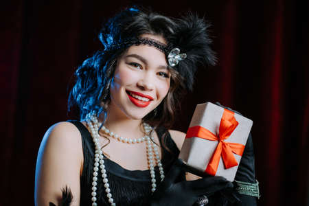 Joyful vintage styled woman dressed in Art Deco era holding gift box with bow on velours background....