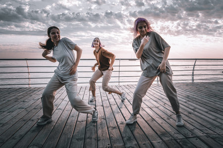 Dancing group of young talented freak women performing freestyle hip-hop moves. females enjoying modern dance expression. Outdoor training near sea or ocean during sunset.の写真素材