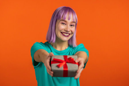 Excited woman with violet hair holding gift box and gives it by hands to camera on orange wall background. Girl smiling, she is happy with present. Studio portraitの写真素材