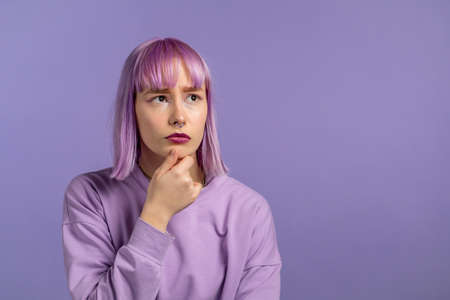 Thinking woman with dyed purple hair looking up on violet studio background. Pensive face...