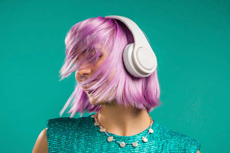 Unusual trendy woman with dyed violet hair listening music, dancing in headphones on turquoise background. Fashionista teen girlの写真素材