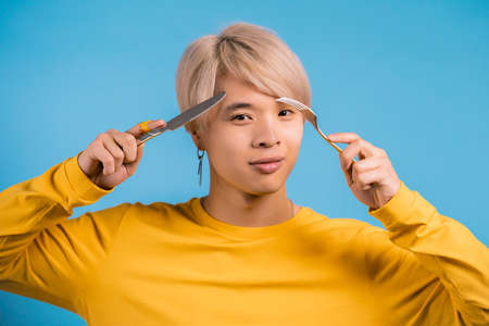Portrait of hungry asian man with fork and knife. Guy waiting for serving dinner dishes with cutlery...