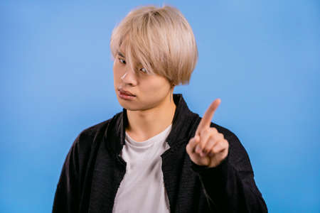 Portrait of serious korean man showing rejecting gesture by stop finger sign. Asian guy with dyed blonde hair isolated on blue background.の写真素材