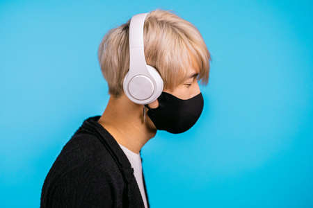 Young man in mask with trendy hairdo with headphones in studio against blue background. Music, radio concept.の写真素材