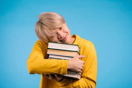 Korean student in yellow wear holds stack of university books from library on blue background in studio. Guy smiles, he is happy to graduate.の写真素材