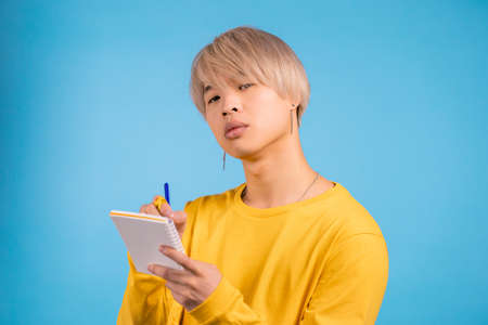 Asian man making notes in planner, korean guy holding pen. He writes future plans and to-do list in notebook for week or month. Keeping personal diary on blue studio background.の写真素材