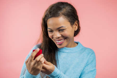 African woman holding small jewelry box with proposal diamond ring on pink wall background. Lady smiling, she is happy to get present, proposition for marriageの写真素材