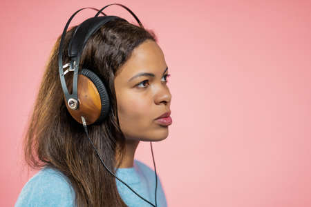 Mixed race woman dancing with wireless blue headphones isolated on pink studio background. Cute lady portrait. Music, radio, happiness, freedom, youth concept.の写真素材