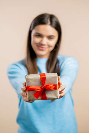 Excited woman holding gift box and gives it by hands to camera on light wall background. Girl smiling, she is happy with present. Studio portraitの写真素材