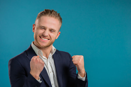 Businessman in corporate suit is very glad and happy, he shows yes gesture of victory, guy achieved result, goals. Surprised excited happy guy on blue backgroundの写真素材