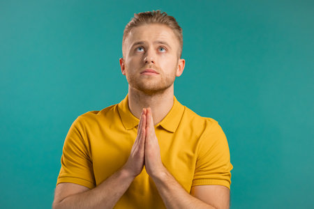 Young man praying over blue background. Guy begging someone satisfy his desires, help with.の写真素材