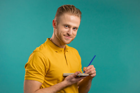 Handsome young man making notes in planner, guy holding pen, reflects on plans. Guy in yellow wear makes records in diary on blue studio background.の写真素材