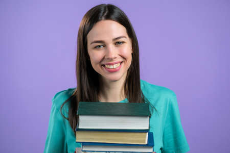 Student holds stack of university books from library on violet background in studio. Woman smiles, she is happy to graduate.の写真素材