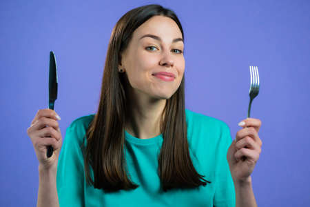 Portrait of hungry woman with fork and knife. Lady waiting for serving dinner dishes with cutlery on violet studio background.の写真素材