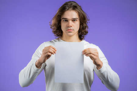 Portrait of young serious man holding white vertical a4 paper isolated on violet studio background.Copy space.の写真素材