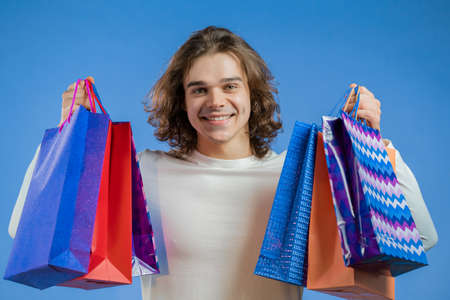 Handsome man with long hairstyle holds shopping paper bags on blue studio background. Guy bought...
