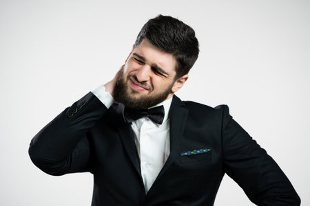 Tired young man having pain - neck ache. Guy in black holiday tuxedo takes break and kneads sore...