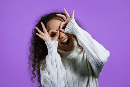 Positive girl making OK sign over eyes like glasses on purple background and smiles to camera. Body...
