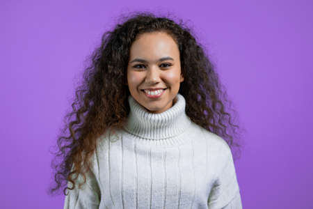 Portrait of pretty woman on violet studio background. Positive young girl with natural curly hair, she in white sweater smiles to camera.の写真素材