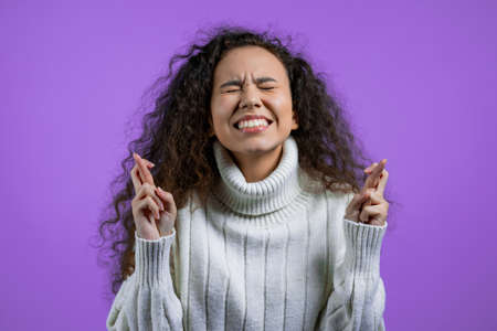 Cute woman in sweater praying with crossed fingers over violet background. Girl begging someone...