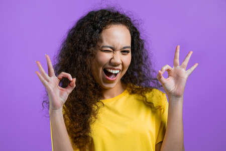 Pretty woman showing ok, okay sign over purple studio background. Positive young girl with natural curly hair smiles to camera. Winner. Success. Body language.の写真素材