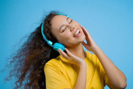 Attractive woman dancing with wireless headphones on blue studio background. Cute girl dancing and smiling. Music, radio, happiness, freedom, youth concept.の写真素材