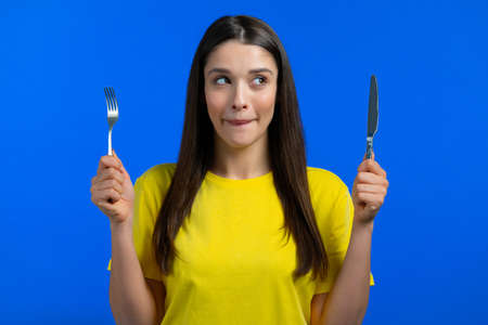 Portrait of hungry woman with fork and knife. Lady with anticipation waiting for serving dinner dishes with cutlery on blue studio background.の写真素材