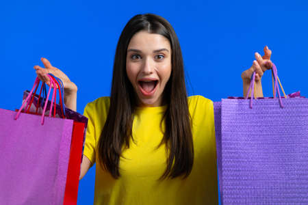 Excited woman with colorful paper bags after shopping on blue studio background. Concept of seasonal sale, purchases, spending money on giftsの写真素材