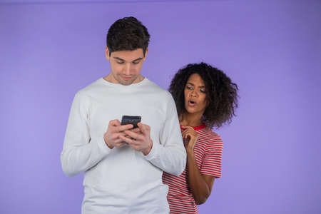 African woman is peeking over boyfriends shoulder, he is communicating via mobile telephone. Girl shocked by jealousy and betrayal of husband. Purple background. Secrets concept, hidden double lifeの写真素材