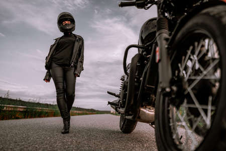 Sexy woman in helmet sits goes to her classic black motorcycle. Female confident motorcyclist alone...
