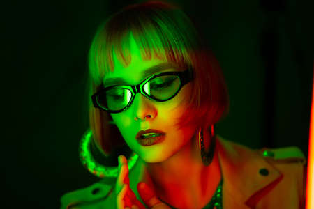 Charming woman with dyed green hair posing under neon light. Unusual mysterious lady in sunglasses, nightlife concept. Modern model pop outfit, influencer lifestyle.の写真素材
