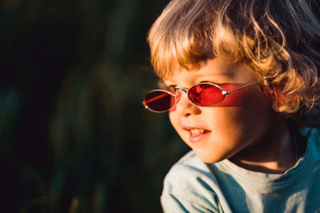 Funny curly hair baby boy in red sunglasses standing in fresh green wheat field. Lovely toddler child explores plants, nature in spring. Childhood, children fashion style conceptの写真素材