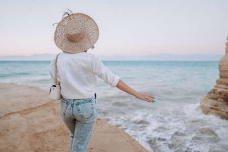 Pretty woman standing on beach near Mediterranean sea. Lady tourist in straw hat watching beautiful blue water surface, nature background. Windy weather, golden hour.の写真素材