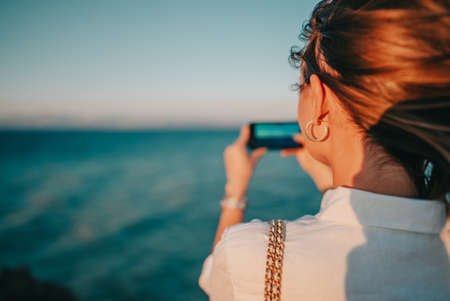 Young woman filming video with mobile phone on beach near blue sea. Girl using smartphone, taking photos for social app, enjoying vacation time.の写真素材