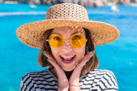 Surprised pretty woman in yellow sunglasses and straw hat says WOW with open mouthed in amazement to camera on teal blue sea background. Lady on luxury travel destination, vacation lifestyle concept.の写真素材