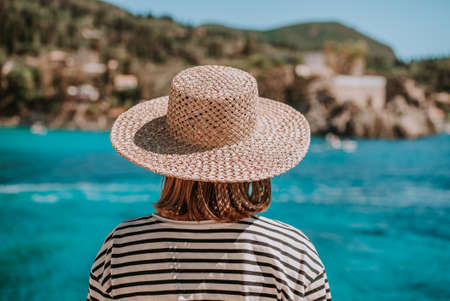 Back view of woman in striped cruise outfit, straw hat. Lady standing on stone beach. Teal sea background. South Europe Riviera, travel destination, luxury vacation concept.の写真素材