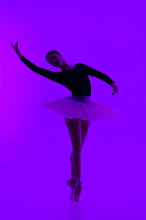 Beautiful ballerina on purple background dancing ballet. Woman performs smooth movements. Sensual dancer in tutu dress on scene under neon light..の写真素材