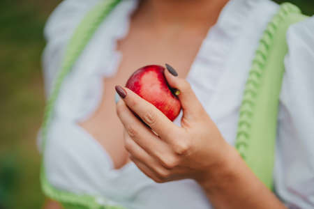 Woman in dirndl offers red apple as symbol of temptation, poison. Lady holding in hand ripe fruit. Fairy tale concept, halloween, cosplay, harvestの写真素材