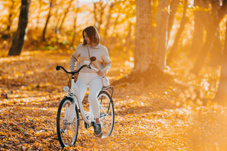 Beautiful portrait of young woman cycling alone in autumn park. Sunny day, golden leaves in forest. Trendy lady on vintage white bicycle, healthy lifestyle, aesthetic sceneの写真素材