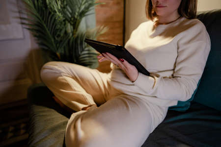 Woman in white reading interesting e-book reader while sitting on couch at home. Cozy winter pic of lady relaxing alone with tablet.の写真素材