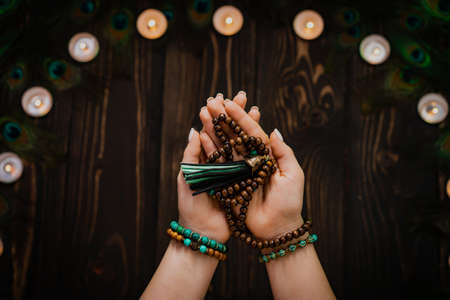Woman holds in hand wooden mala beads strands used for keeping count during mantra meditations. Weaving and creation. Wooden background with candles and feathers. Spirituality, religion, God concept.の写真素材