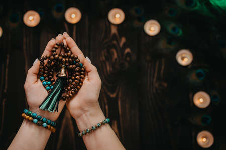 Woman holds in hand wooden mala beads strands used for keeping count during mantra meditations. Weaving and creation. Wooden background with candles and feathers. Spirituality, religion, God concept.の写真素材