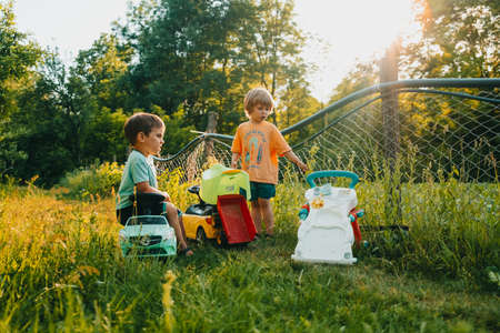 Cute little boys playing together on green summer garden background. Brothers riding plastic colorful cars. Family, happy childhood, friendship concept.の写真素材