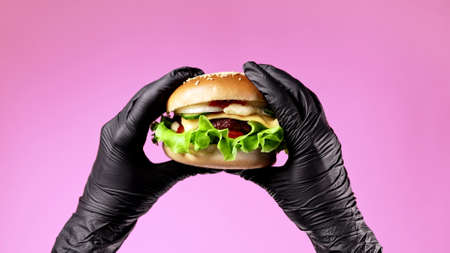 Hands in black latex gloves with burger. Woman holding hamburger. Big appetizing patty with meat cutlet, vegetables, cheese. Fast food concept. Pink background.の写真素材